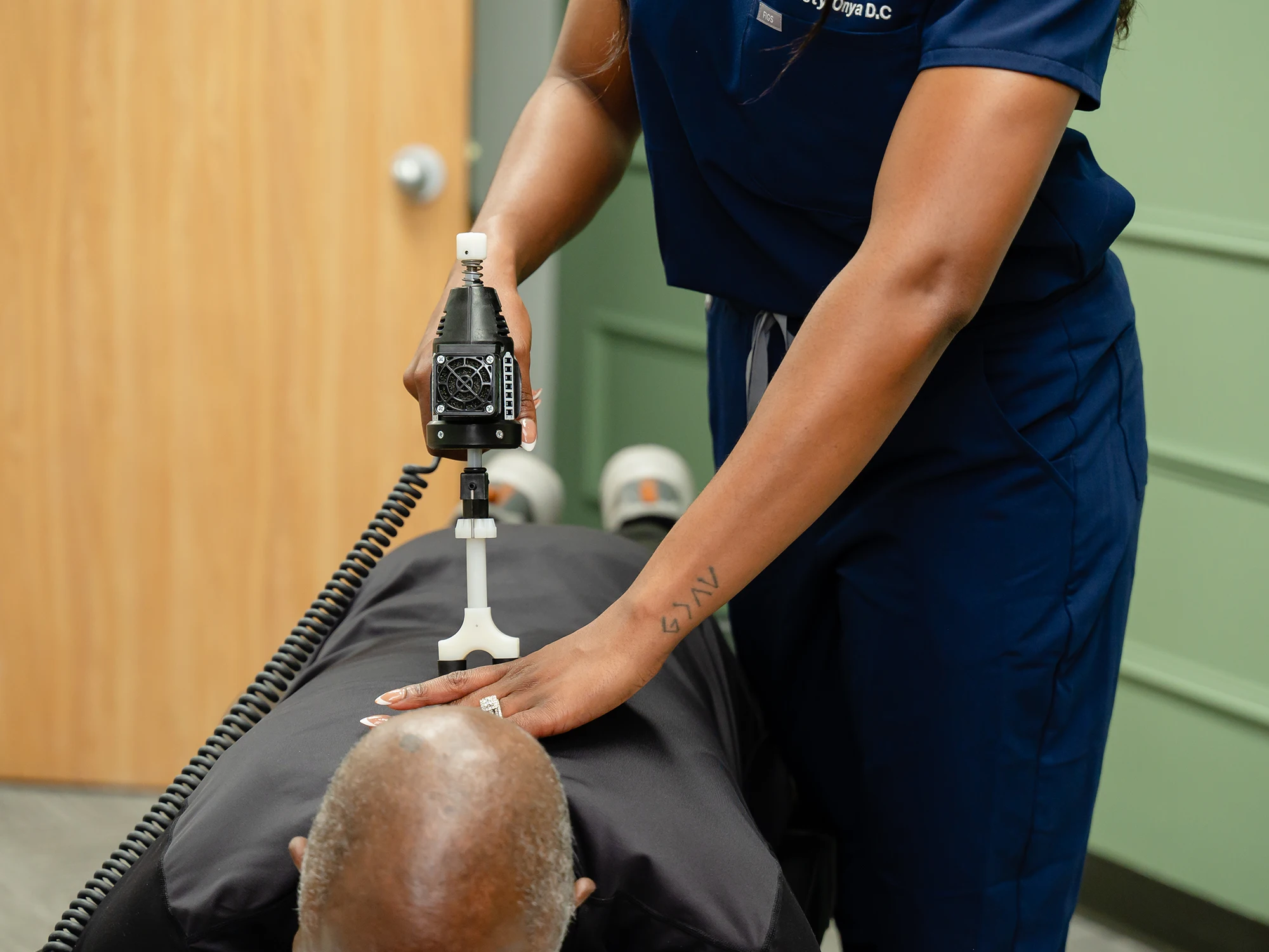 Chiropractor Christy Onya applies a chiropractic adjustment instrument to a patient’s upper back while he lies face down.